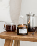 Candle in a jar on a wooden surface with a teapot and glass container in the background.