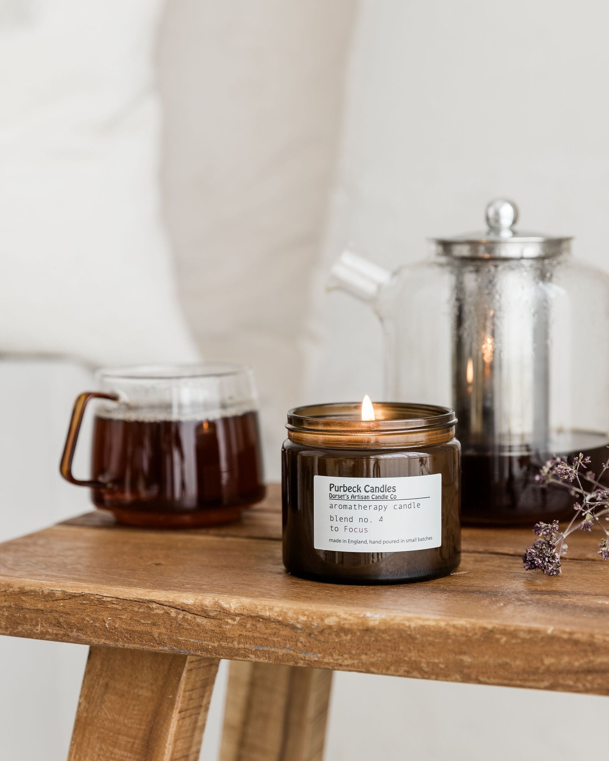 Candle in a jar on a wooden surface with a teapot and glass container in the background.