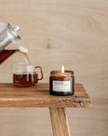 Tea being poured into a glass mug next to a lit candle on a wooden surface.
