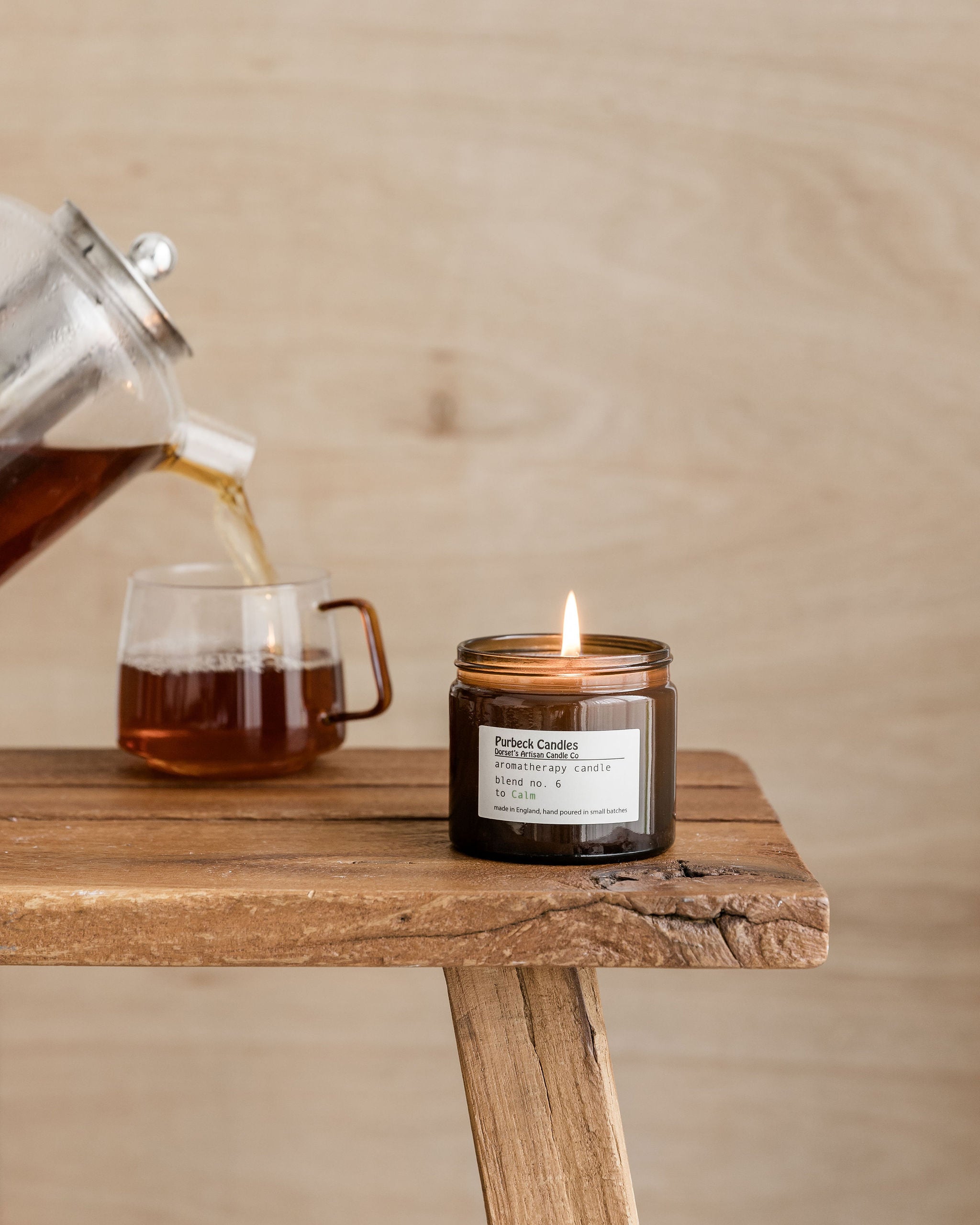 Tea being poured into a glass mug next to a lit candle on a wooden surface.