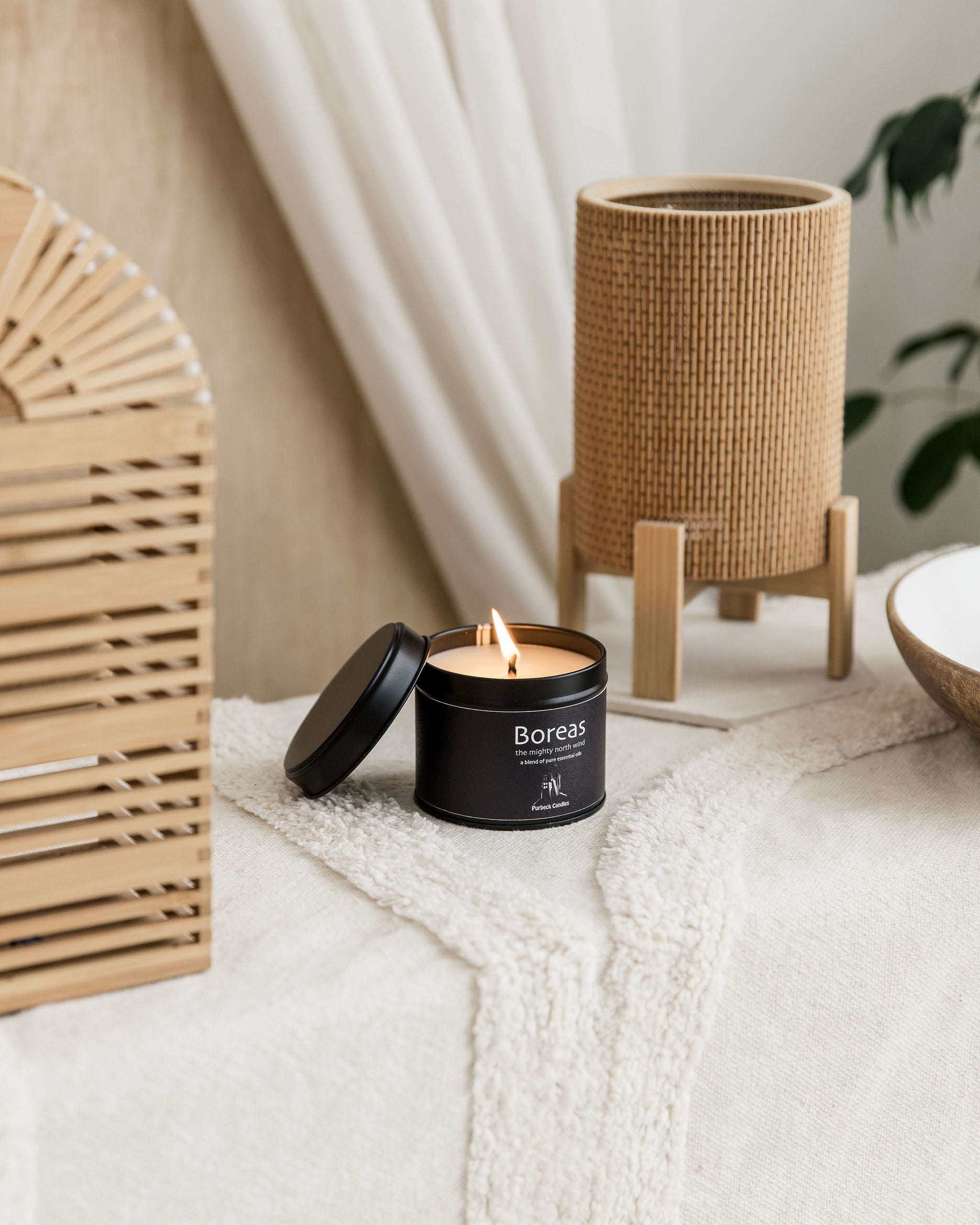 Boreas candle in a black container with a wicker lantern and plant in the background.
