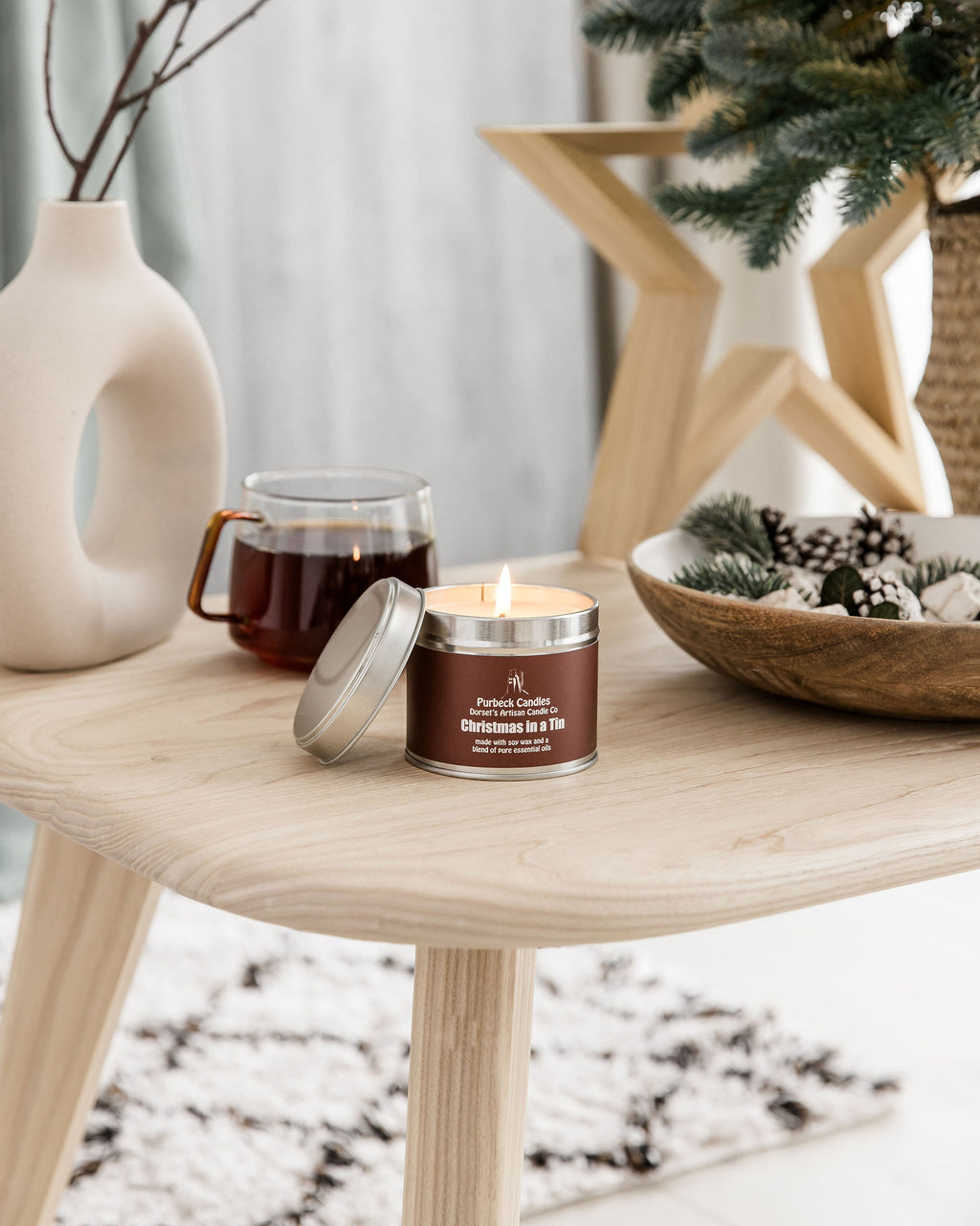 Candle in a tin with a wooden lid on a table next to a mug of coffee and decorative items.