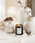 Candle labeled 'cosy' with decorative vases and plants on a soft surface