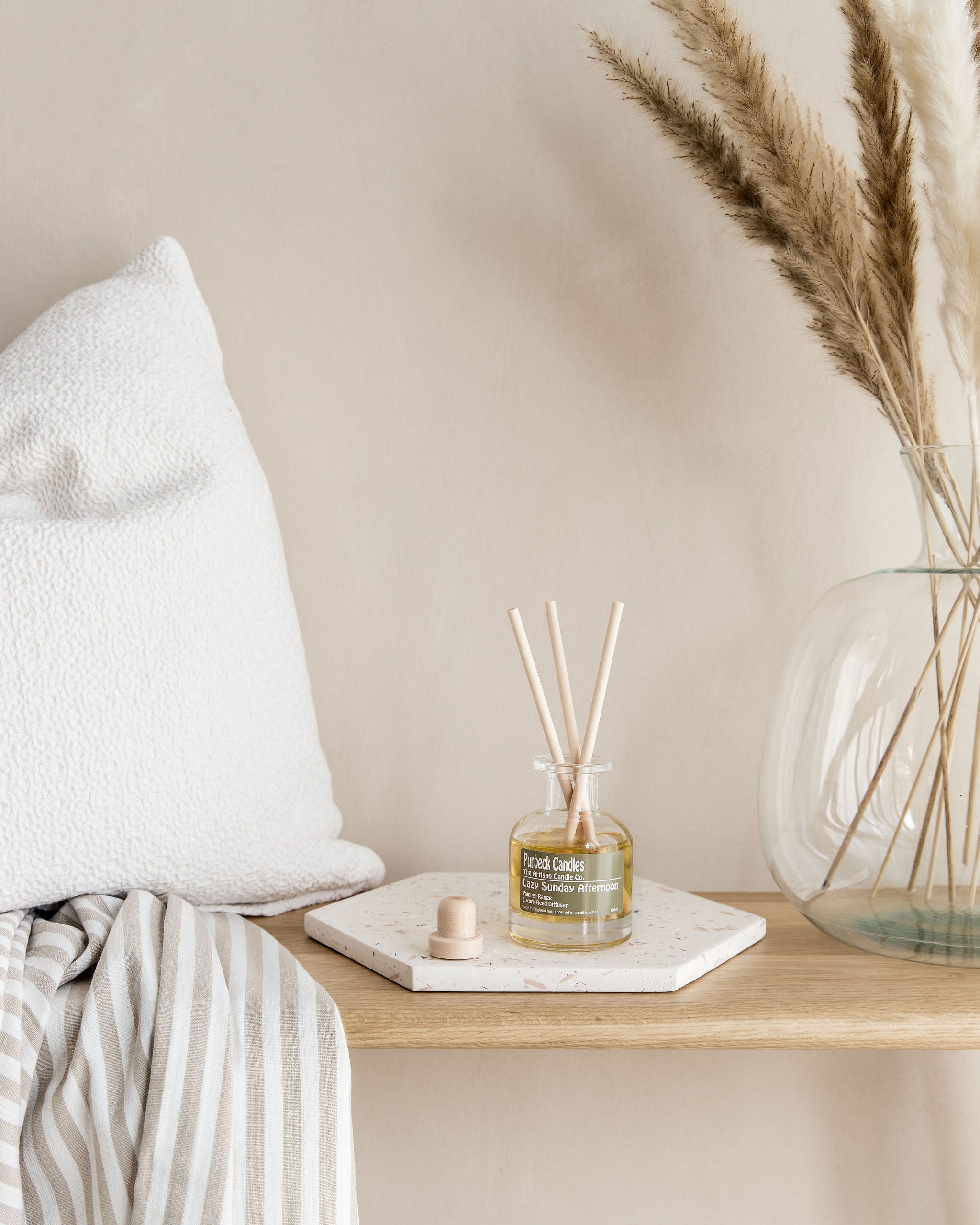 Decorative diffuser with reeds on a marble coaster next to a vase with dried plants on a wooden shelf.