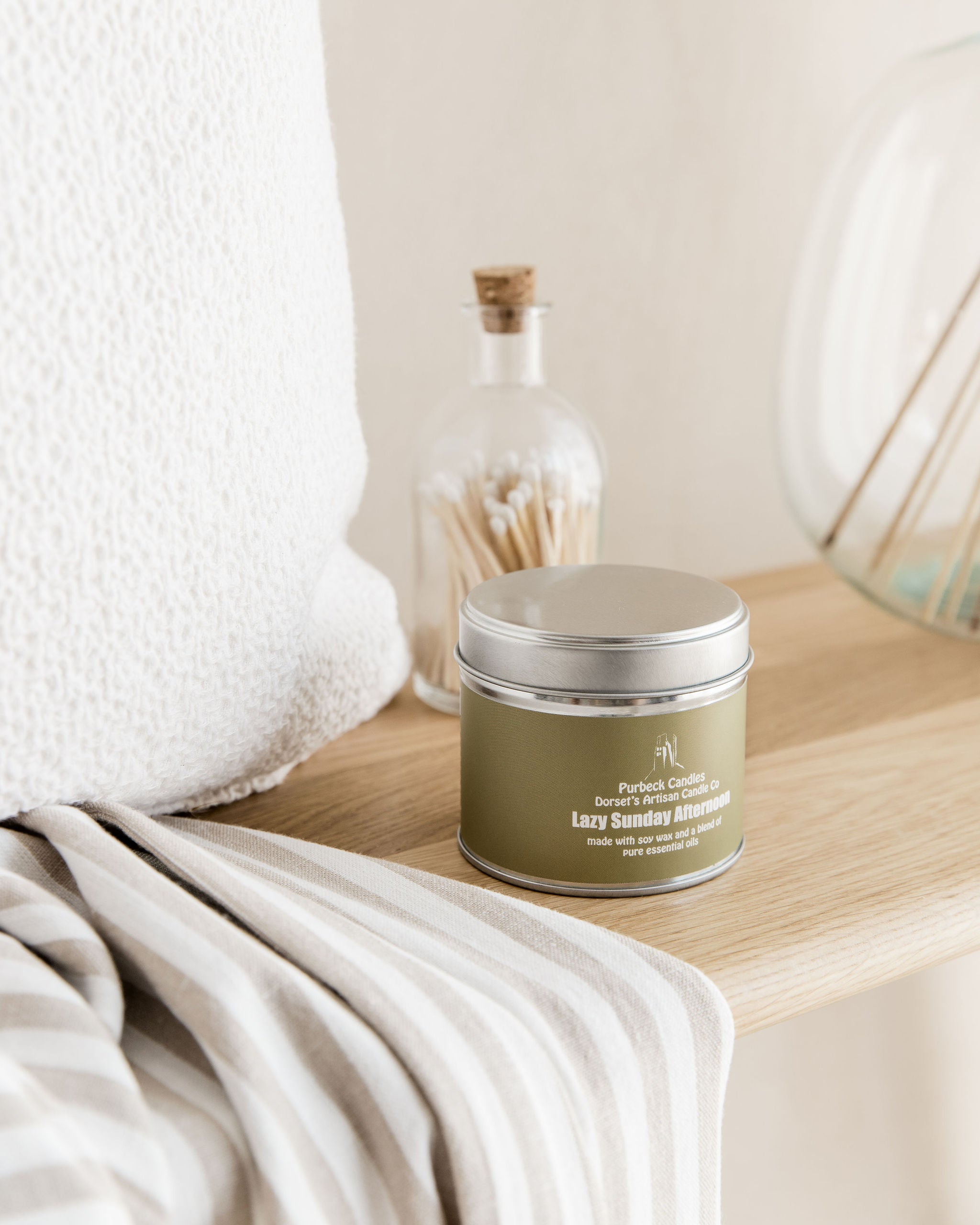 Green candle in a metal tin on a wooden surface with a white towel and decorative items in the background.