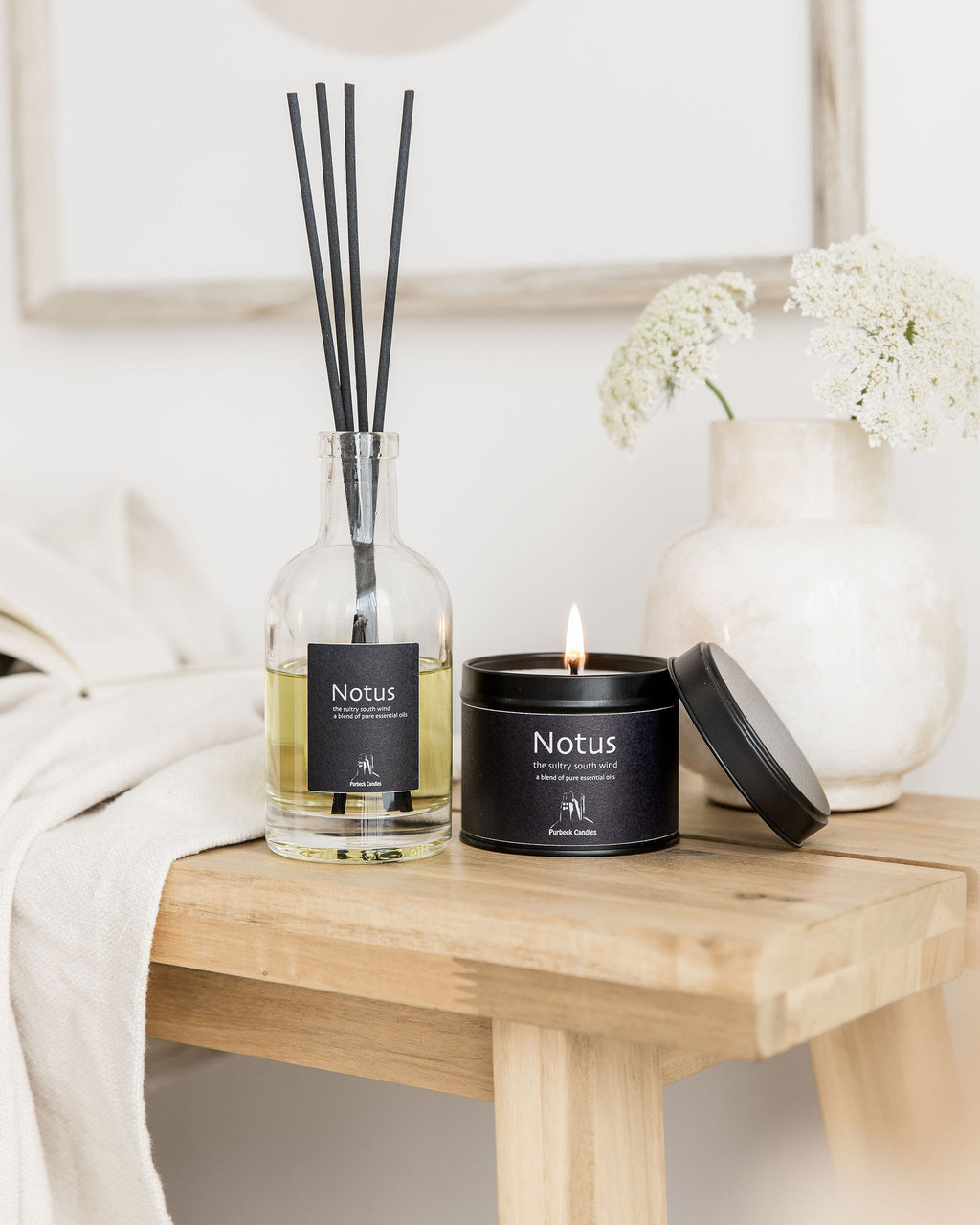 Notus diffuser, candle, and reed sticks on a wooden table with a white vase and flowers in the background.