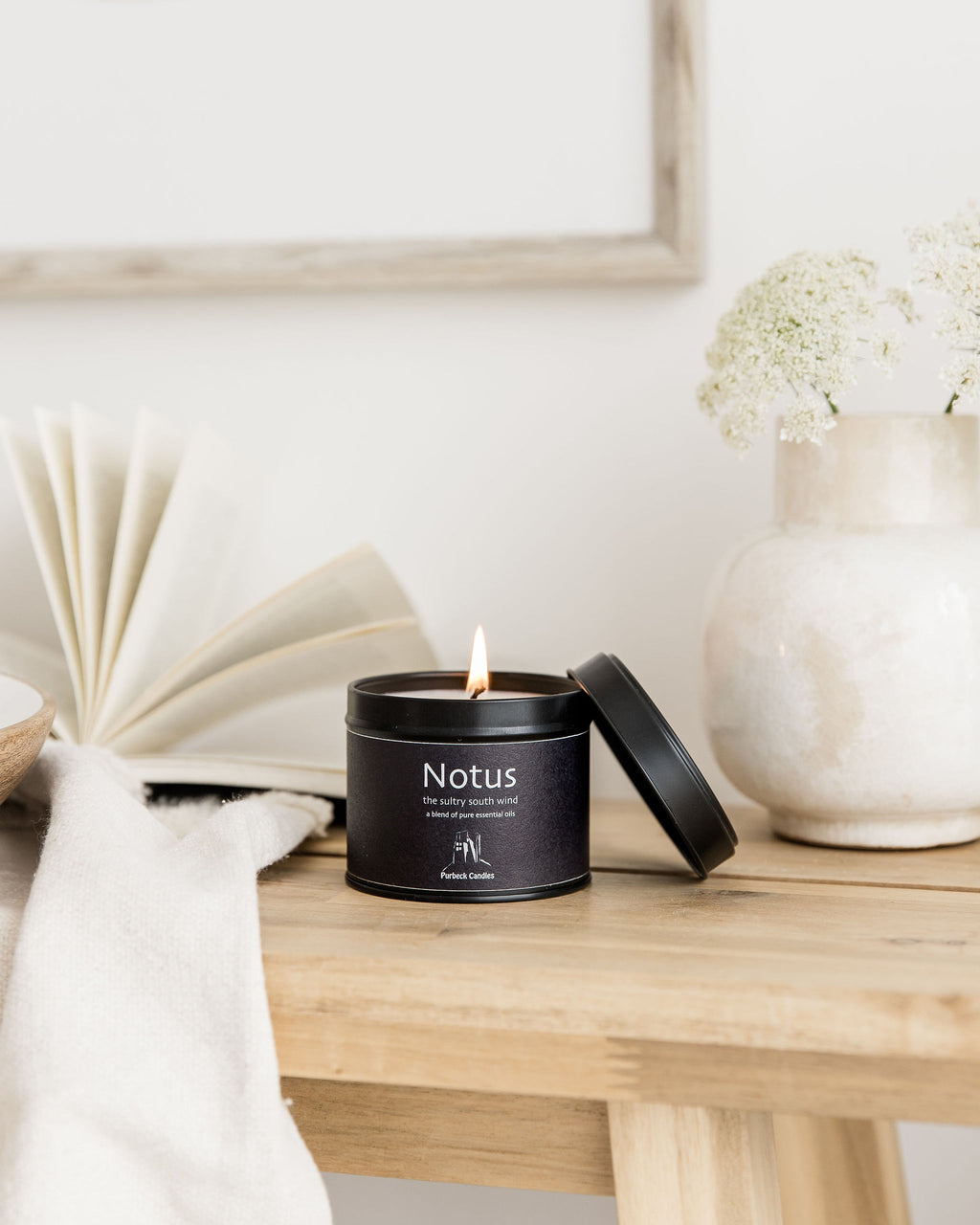Candle in a black container labeled 'Notus' on a wooden surface with a vase and book in the background.
