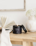 Candle in a black container labeled 'Notus' on a wooden surface with a vase and book in the background.