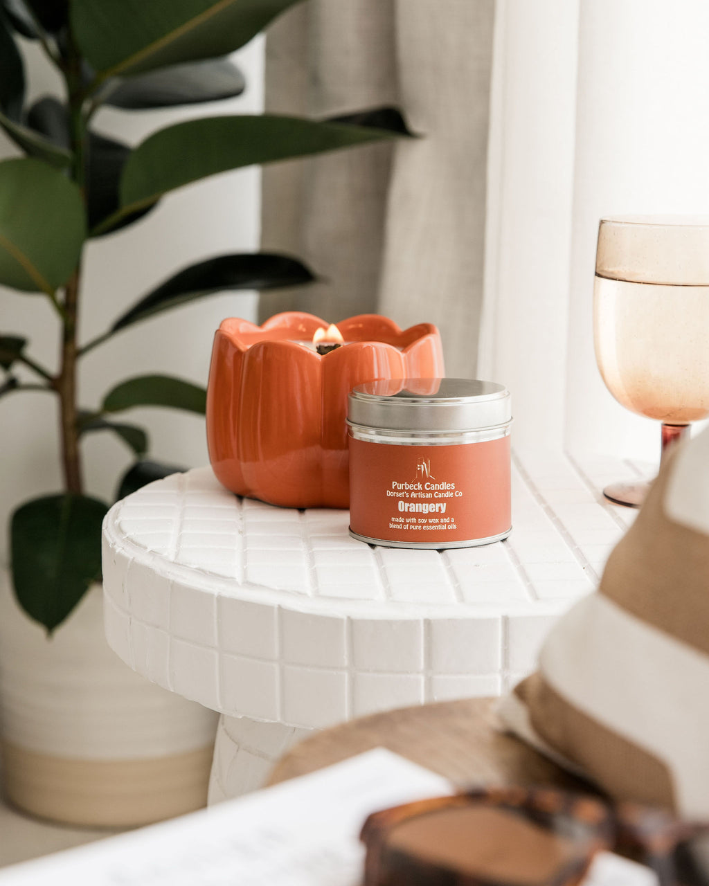 Candle and container on a small table with a plant and glass in the background