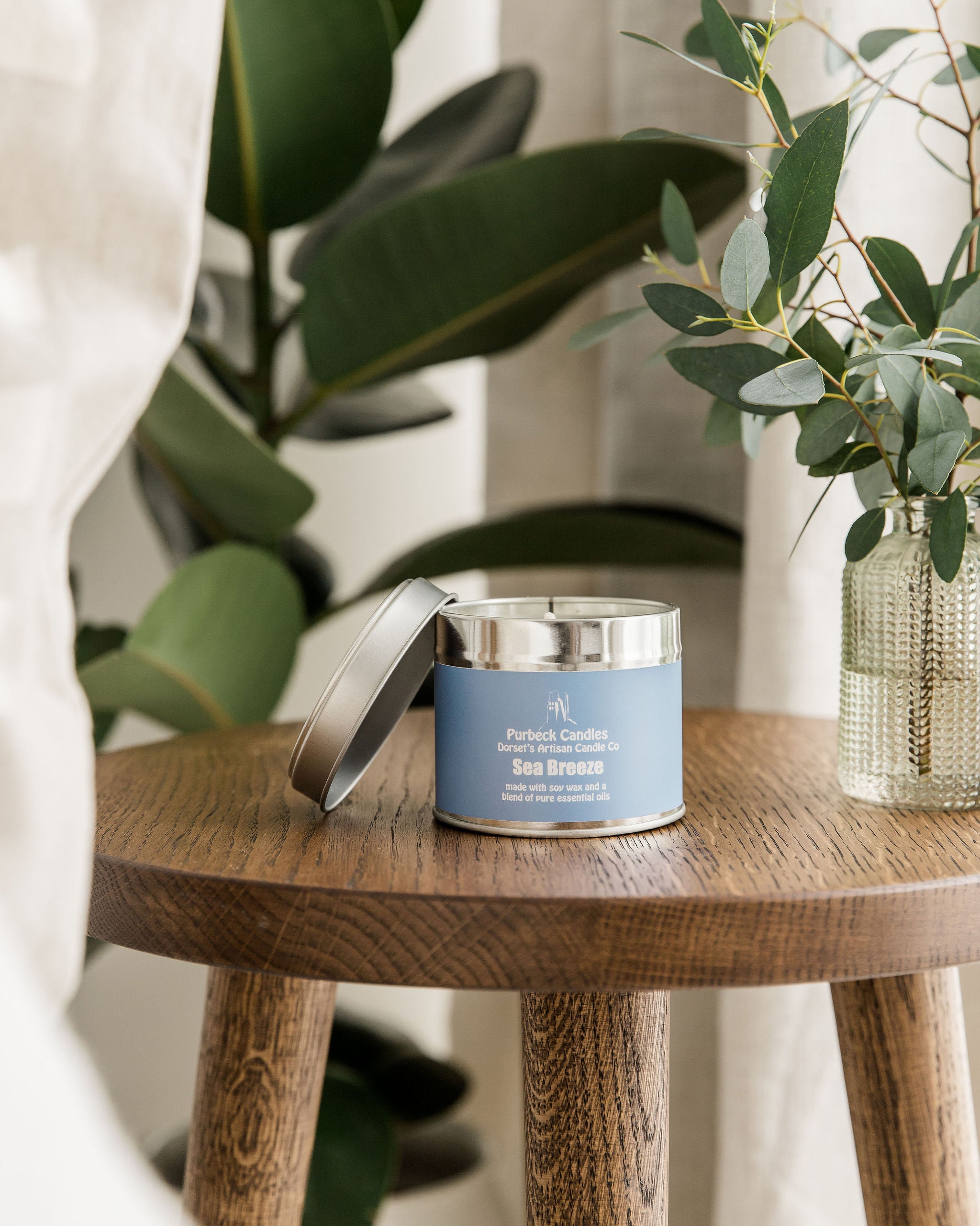 Candle tin with blue label on a wooden table with plants in the background
